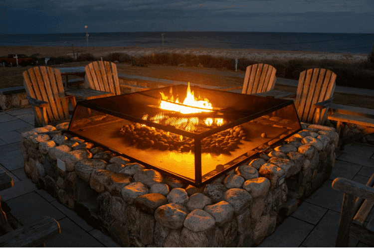 A large stone fire pit with a glass guard surrounded by wooden chairs overlooking the ocean at dusk.