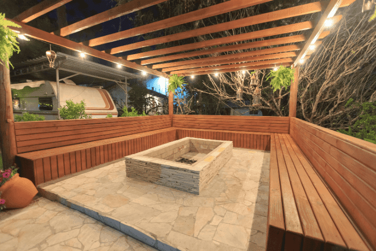 A modern stone fire pit centered within a wooden pergola and bench seating area at night.