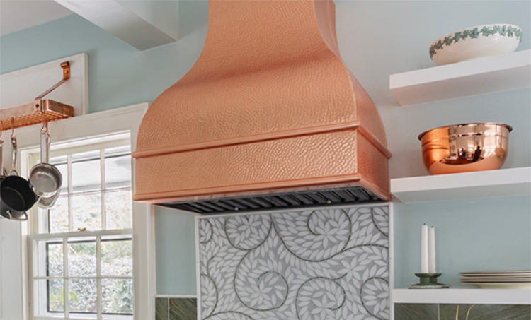 Hammered copper range hood above a stove with a leaf-patterned backsplash and open shelves holding bowls.