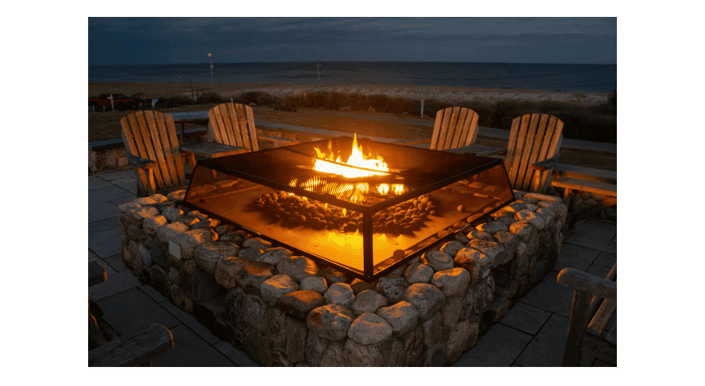 A large stone fire pit with a glass guard surrounded by wooden chairs overlooking the ocean at dusk.