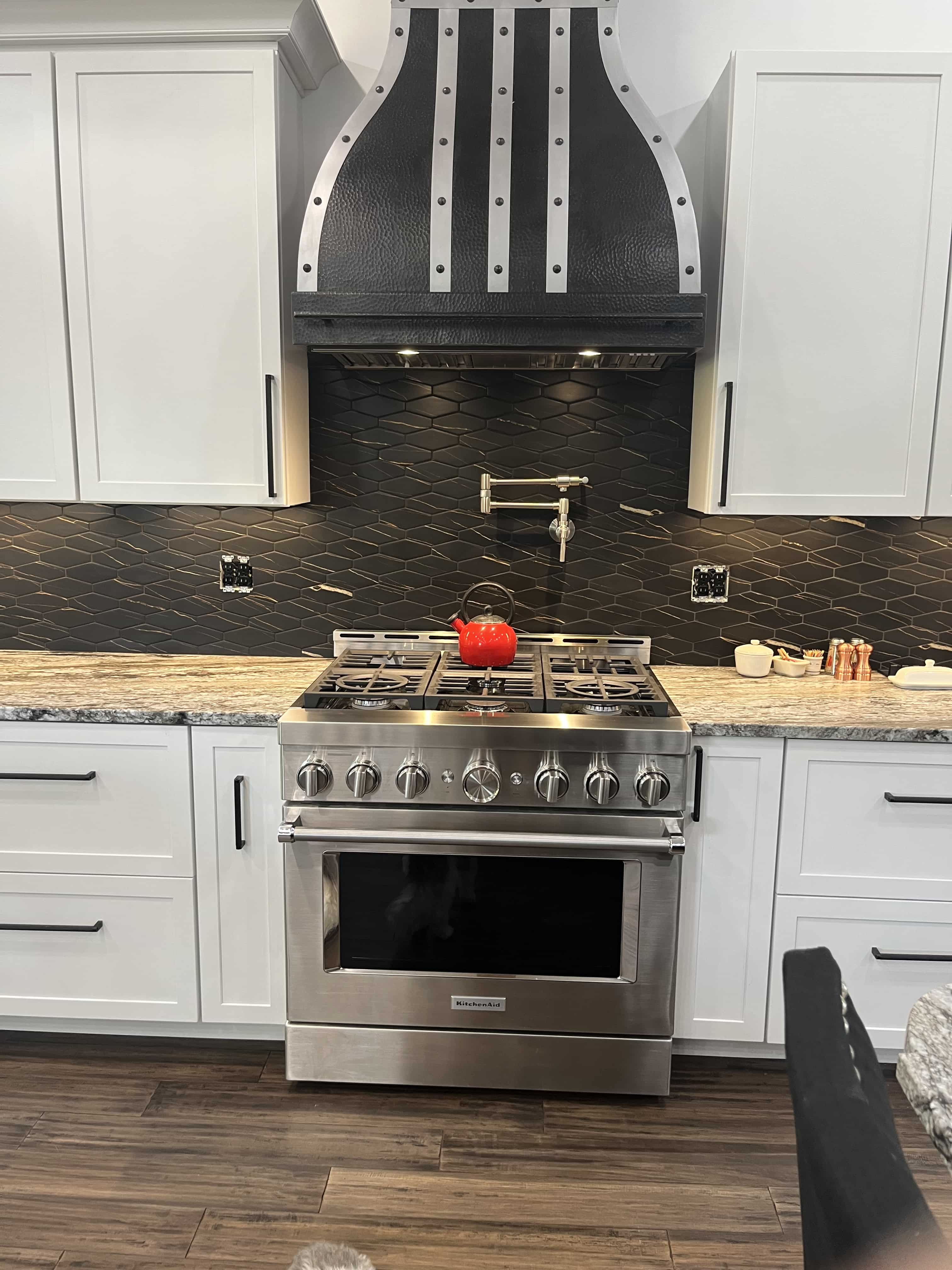 CopperSmith Artisan AT1 Blackened Brass Kitchen Range Hood in a traditional kitchen with white cabinets marble countertops and black backsplash