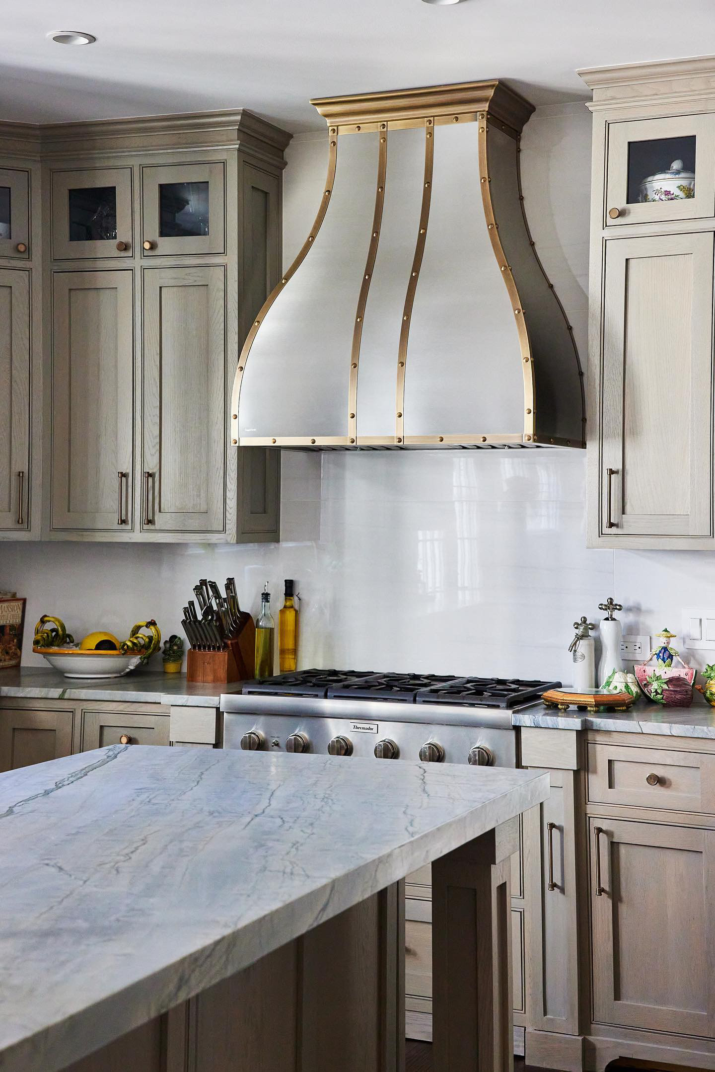 CopperSmith Artisan AT1 Wall Mount Brushed Stainless Steel Range Hood with Light Antique Brass Straps and Rivets in a french style kitchen with marble countertops wood cabinets and white tile backsplash