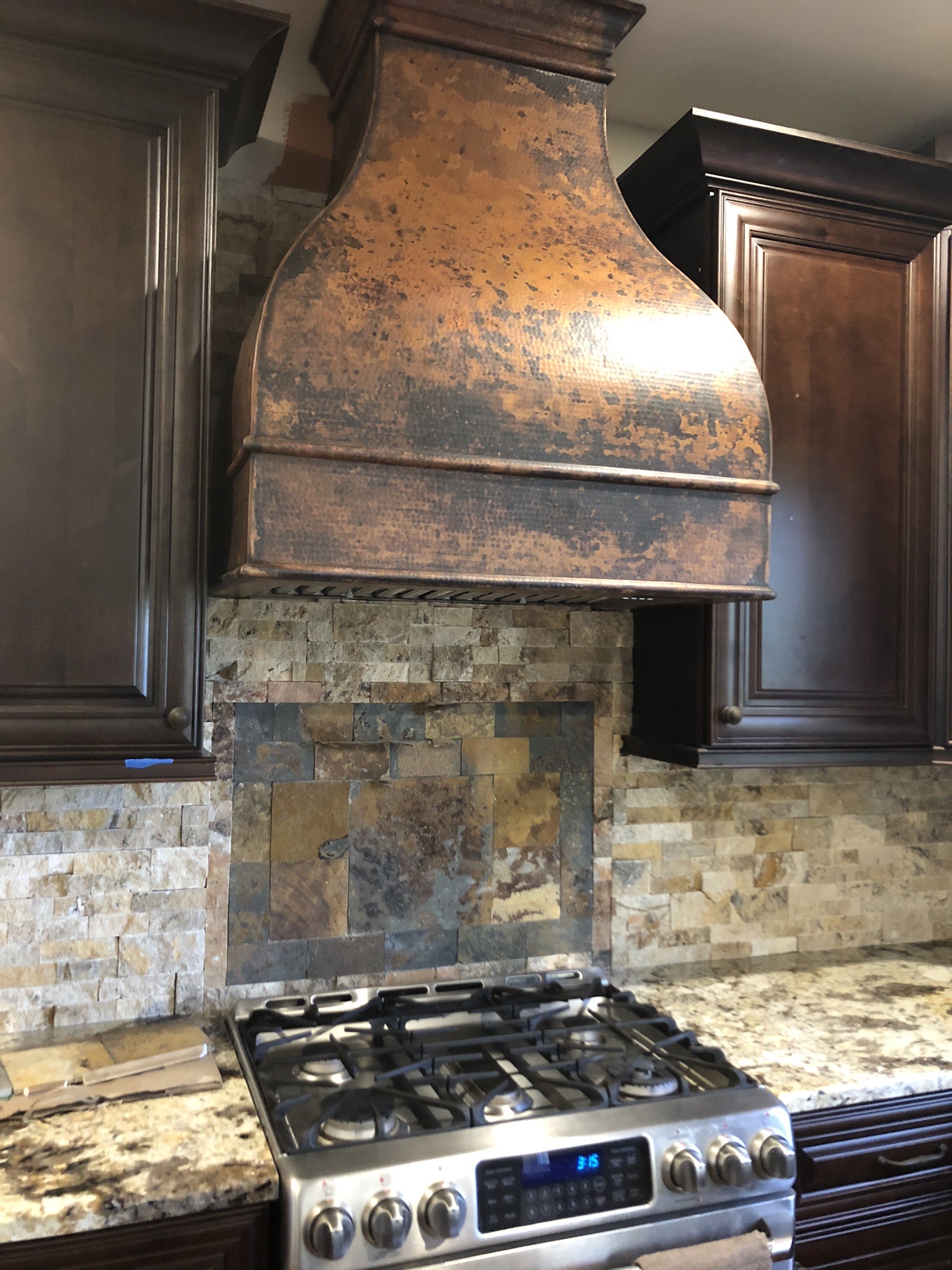 A rustic-inspired kitchen with brown cabinets, marble countertops, brick backsplash, complemented by a stylish range hood design