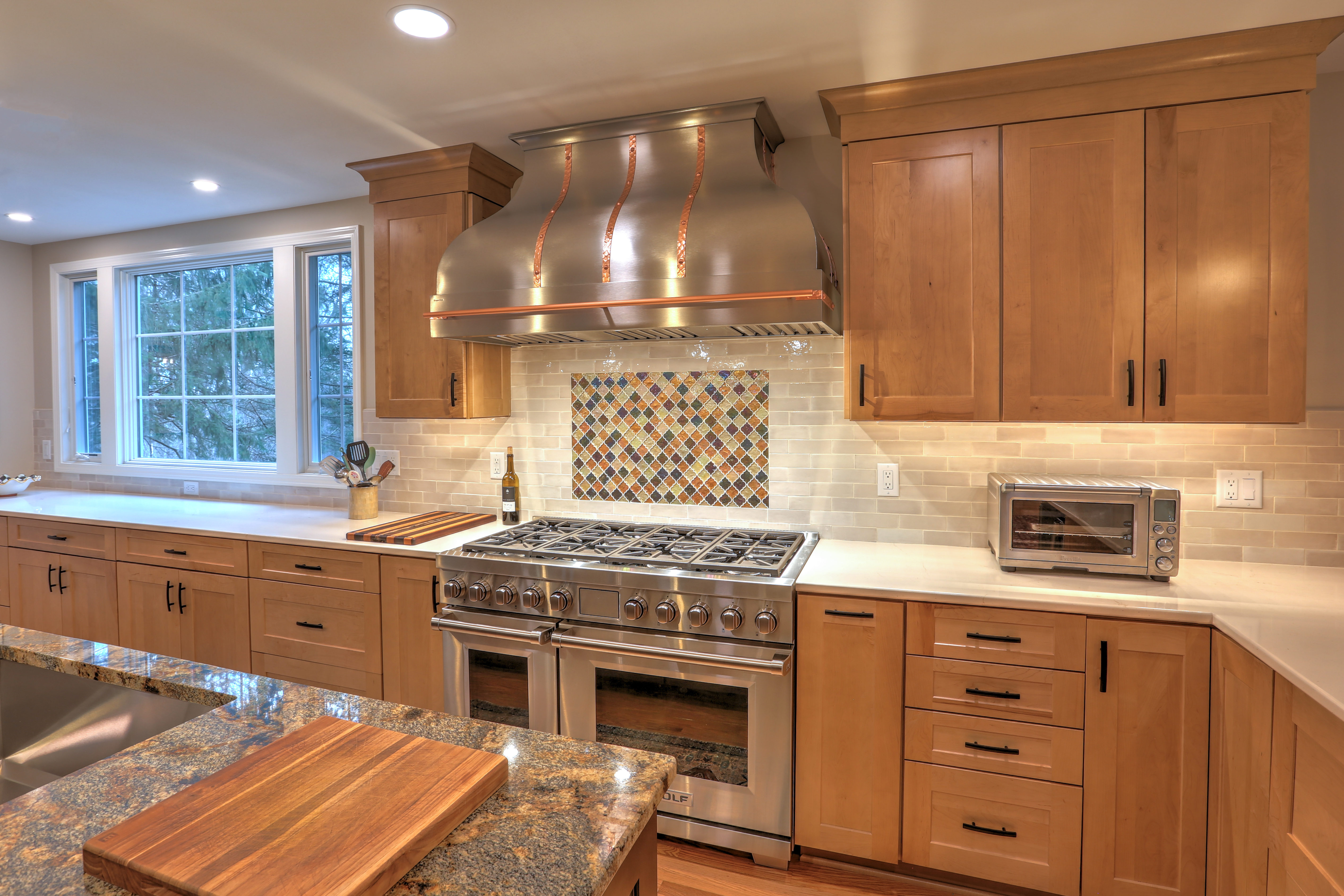CopperSmith Artisan AT3 Wall Mount Stainless Steel Range Hood with Straps and Pot Rail in a rustic kitchen with white tile backsplash, white countertops and wood cabinets