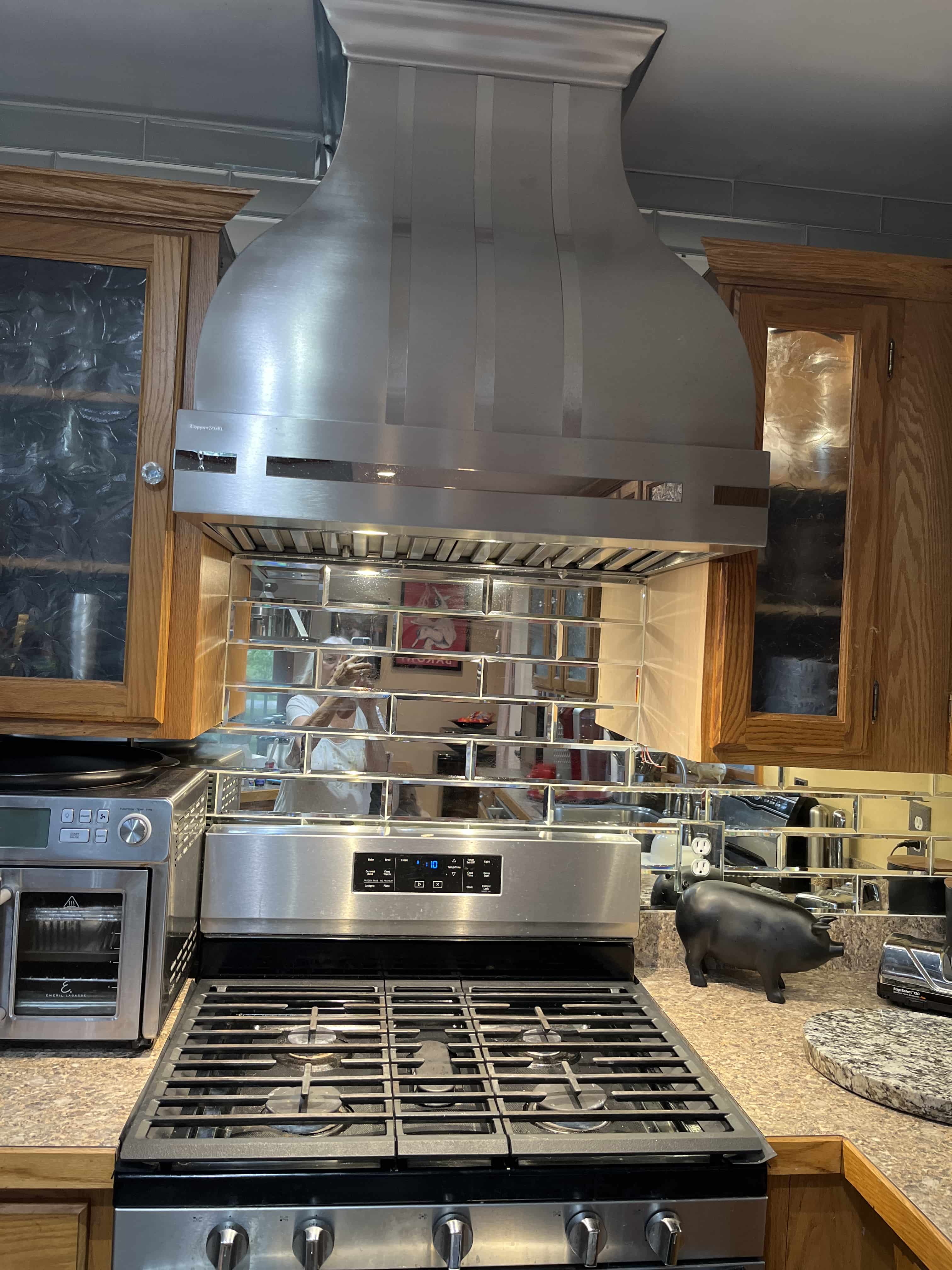 CopperSmith Artisan AT3 Wall Mount Brushed Stainless Steel Range Hood with Polished Stainless Steel Straps in a rustic style kitchen with wood cabinets brown countertops and metal backsplash