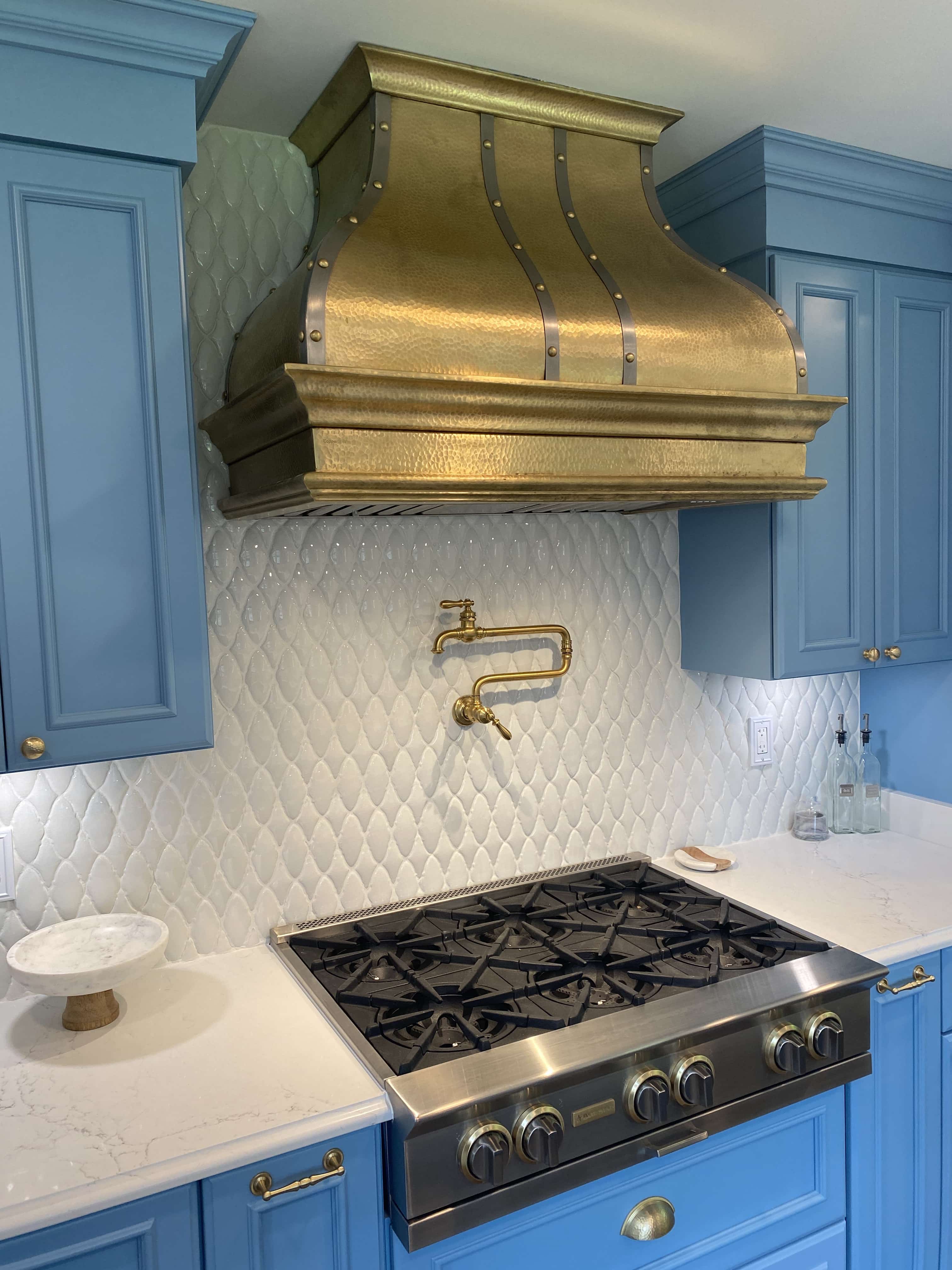 CopperSmith Artisan AT7 Burnished Brass Wall Mount Kitchen Range Hood in a Costal Style kitcvhen with Light Blue cabinets white countertop and white tile backsplash