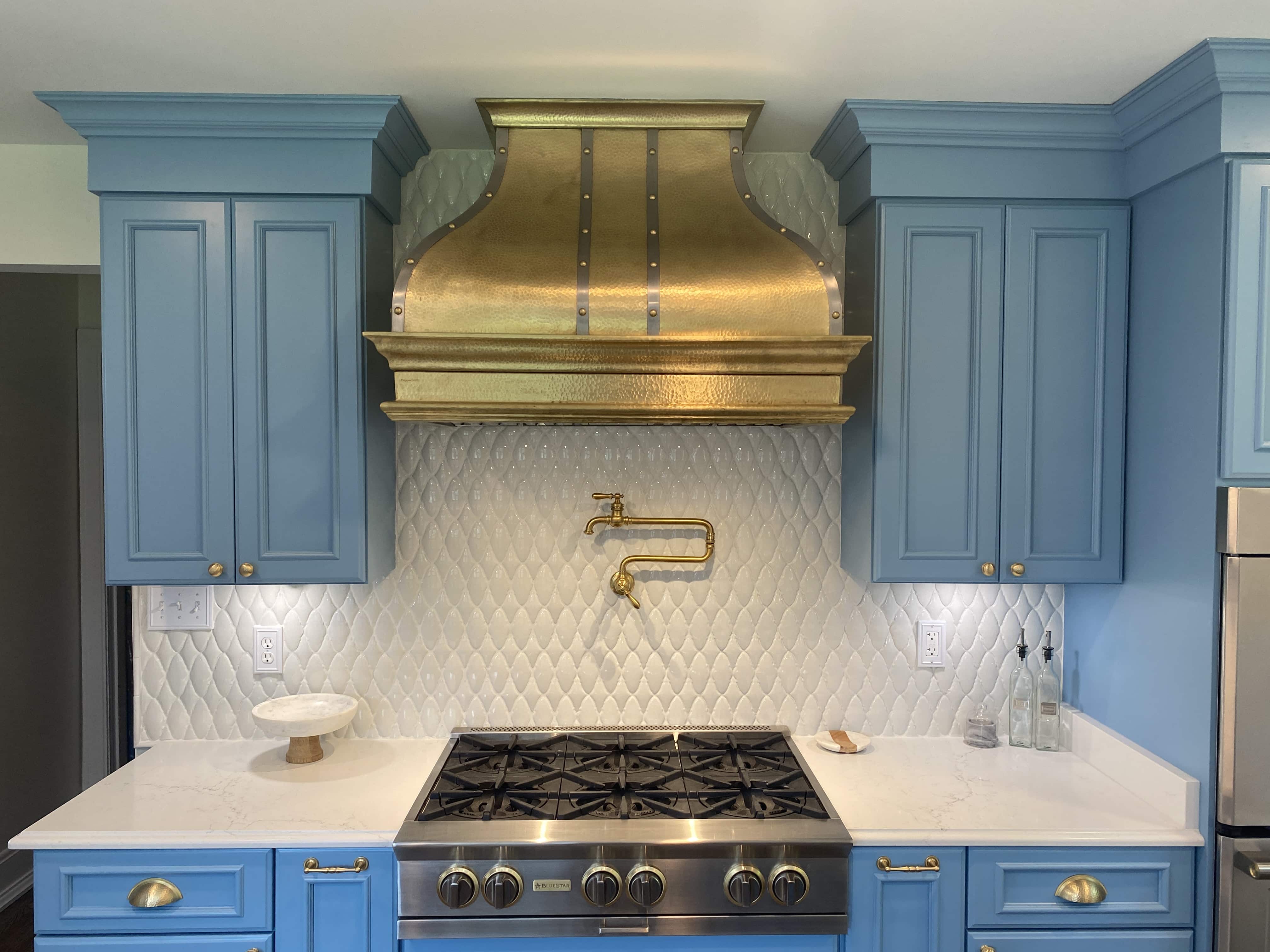 CopperSmith Artisan AT7 Burnished Brass Wall Mount Kitchen Range Hood in a Costal Style kitcvhen with Light Blue cabinets white countertop and white tile backsplash