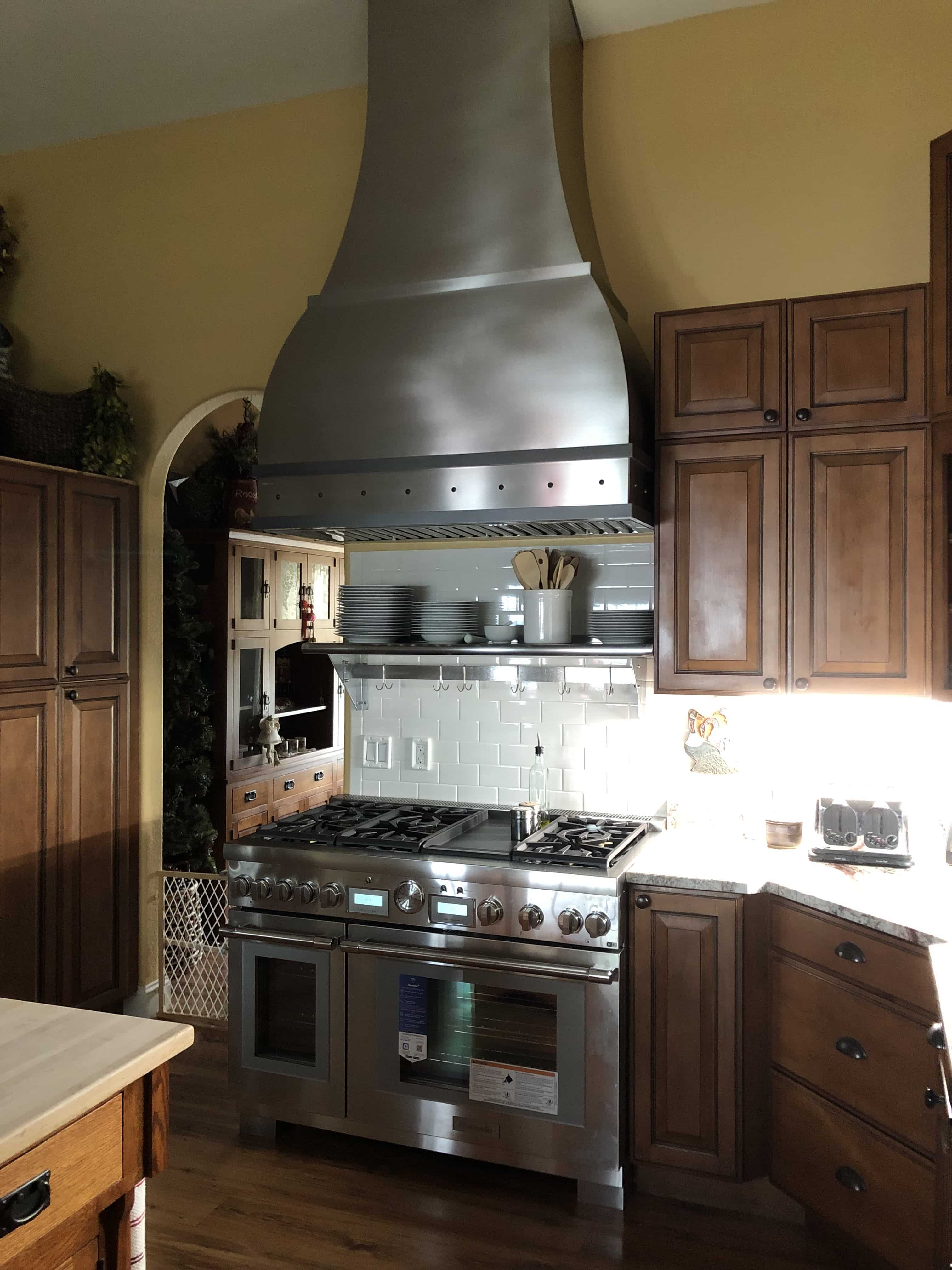 CopperSmith Custom Range Hood Brushed Stainless Steel in a  rustic style kitchen with white tile backsplash white countertops and wood cabinets