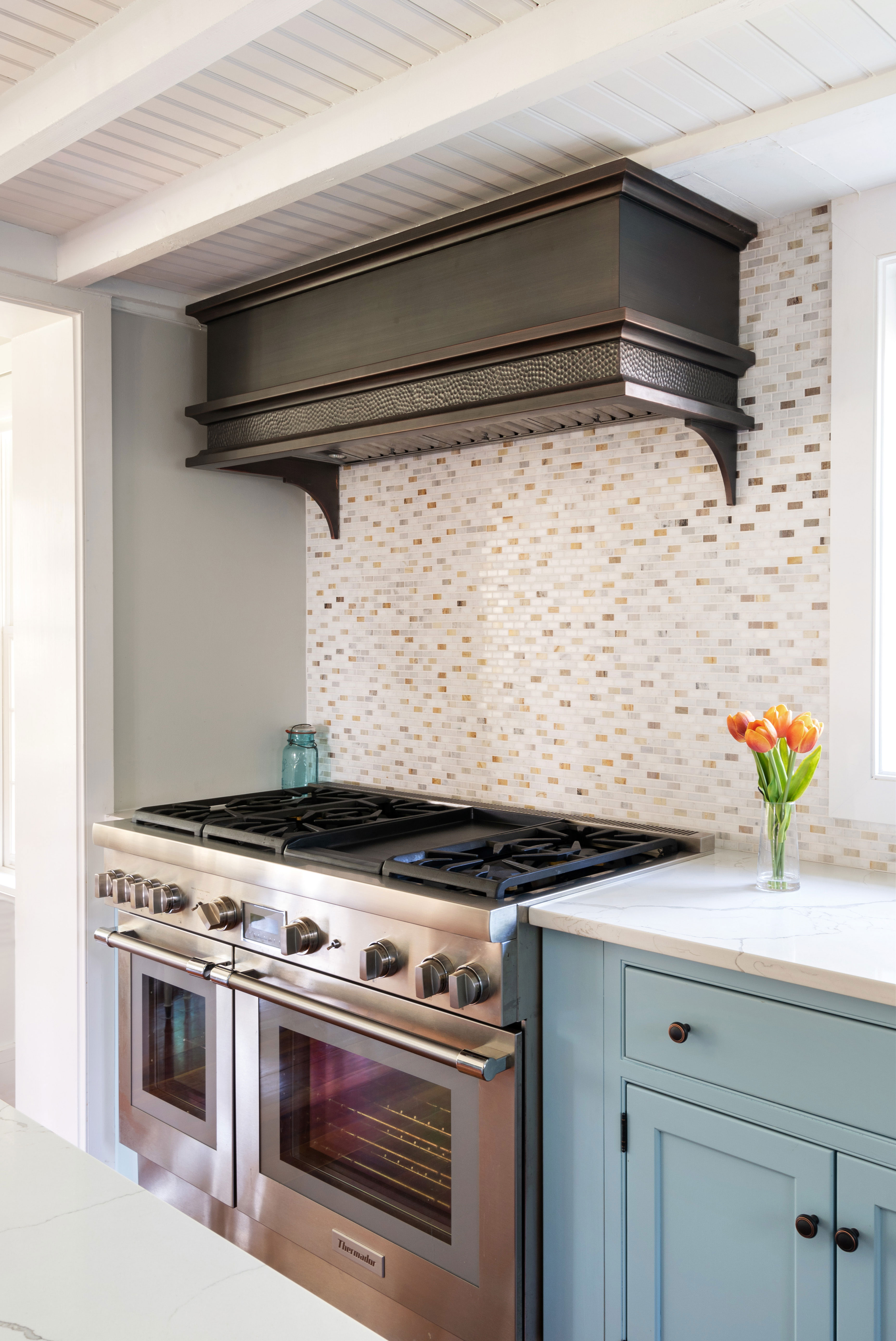 Copper range hood with dappled pattern and brushed finish in a kitchen with wood beam ceilings.