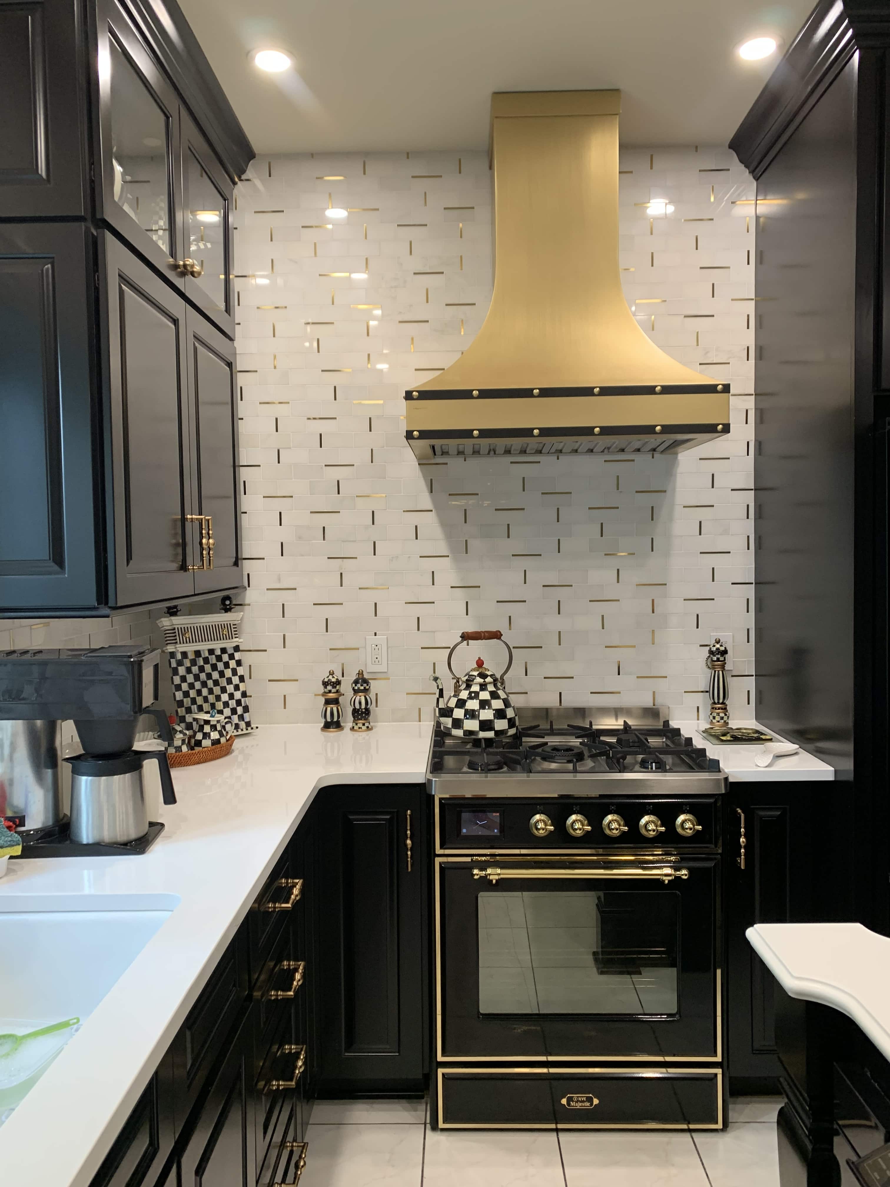 CopperSmith Euro ES3 Matte Brass Kitchen Range Hood with Black Enamel Straps in cottage style features black cabinets white countertops and white tile backsplash