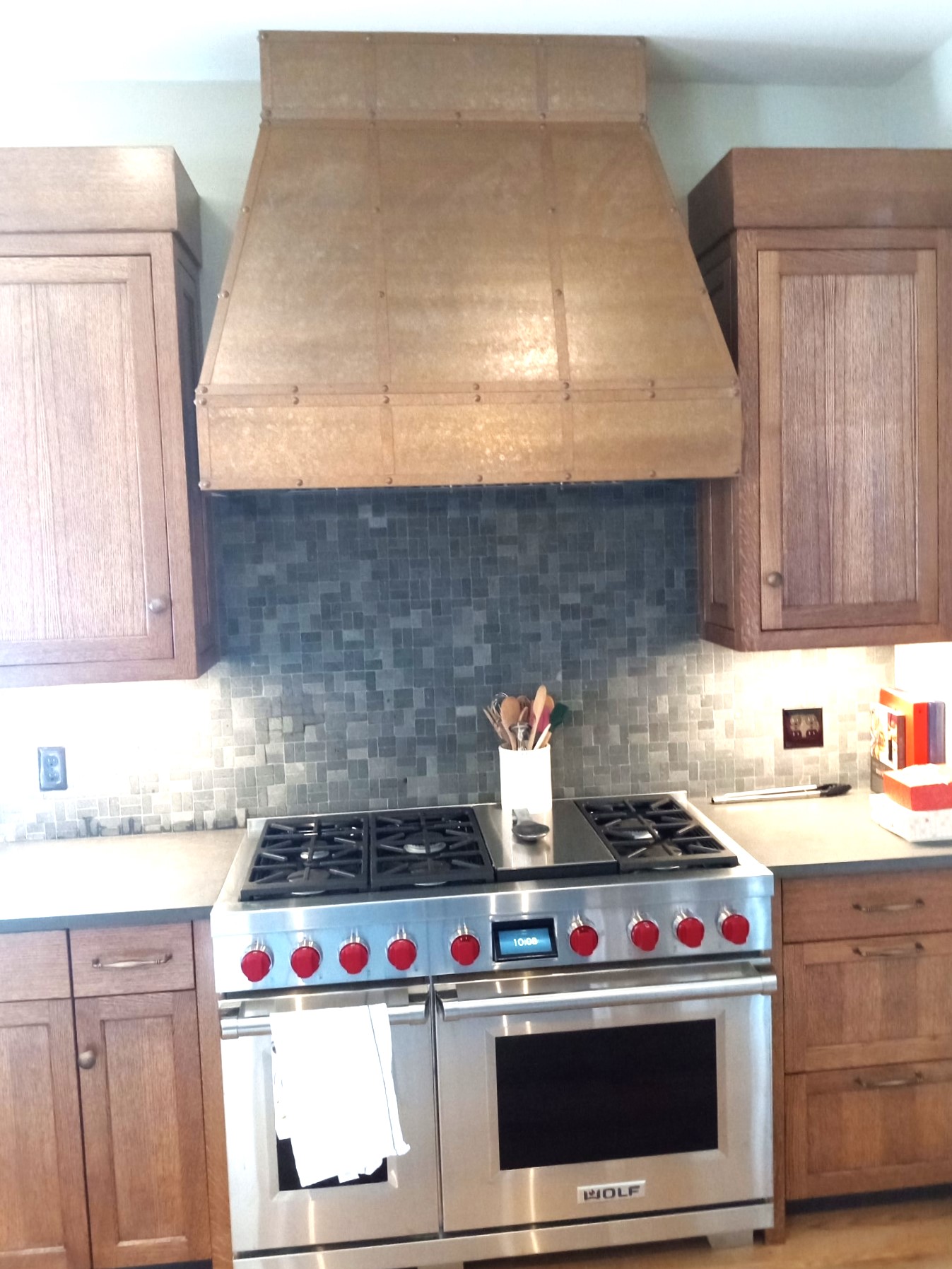 CopperSmith Modern MX3 Wall Mount Weathered Copper Range Hood with Straps and Rivets in a traditional kitchen with brick backsplash wood cabites and marble countertops