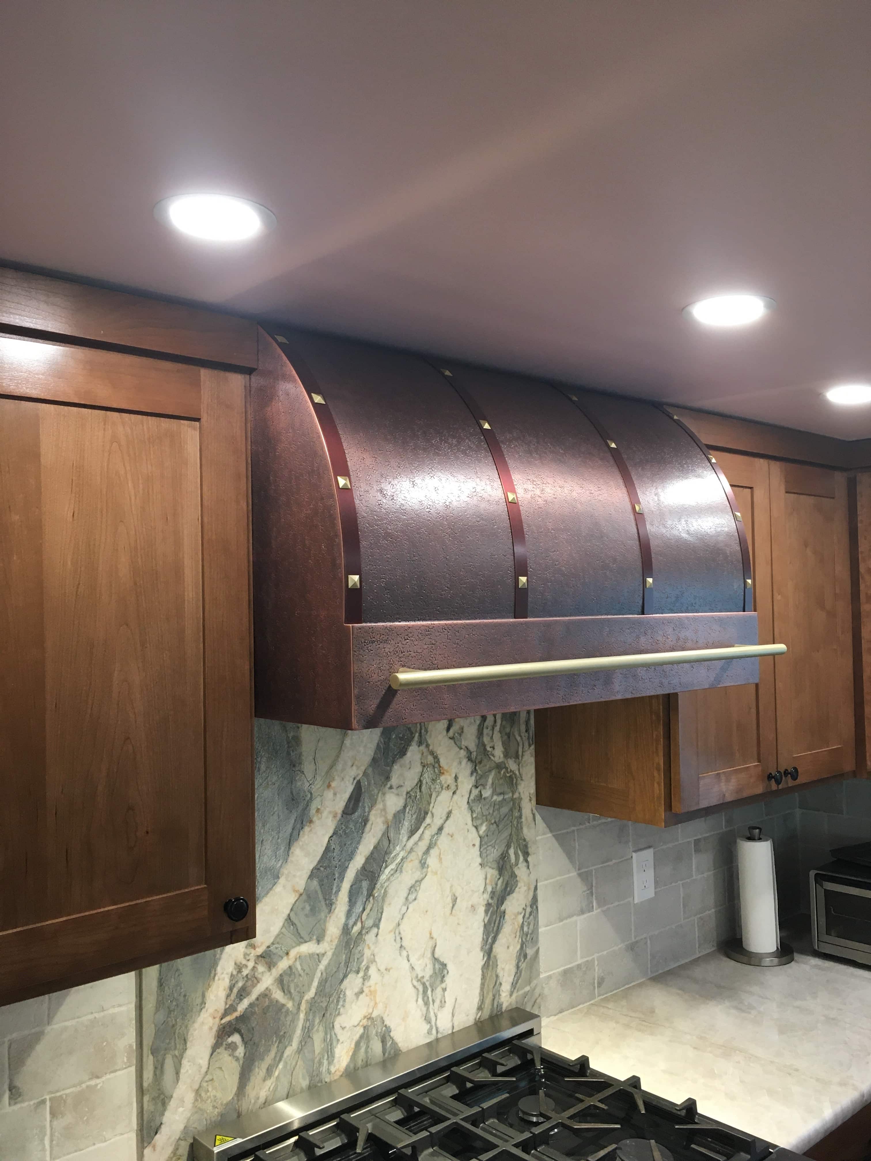 CopperSmith Premier PR4 Light Antique Copper Kitchen Range Hood with Rose Copper Straps Matte Brass Rivets and Pot Rail features wood cabinetry and marble backsplash