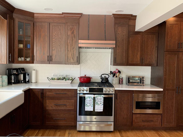 A craftsman kitchen with brown cabinets, white countertops, brick backsplash is accentuated by a stylish range hood