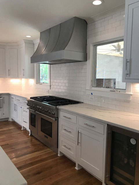 Contemporary kitchen remodel featuring white kitchen cabinets and marble countertops is enhanced with aluminum range hood and stylish brick backsplash