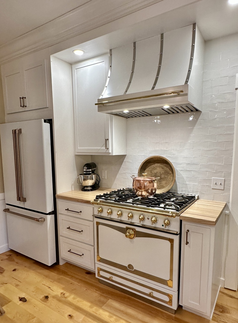 CopperSmith Premier PR7 Wall Mount RAL 9003 Signal White Semi Gloss Range Hood with Brushed Stainless Steel Straps and Polished Brass Rivets in a french style kitchen with white backsplash white cabinets and wood countertops