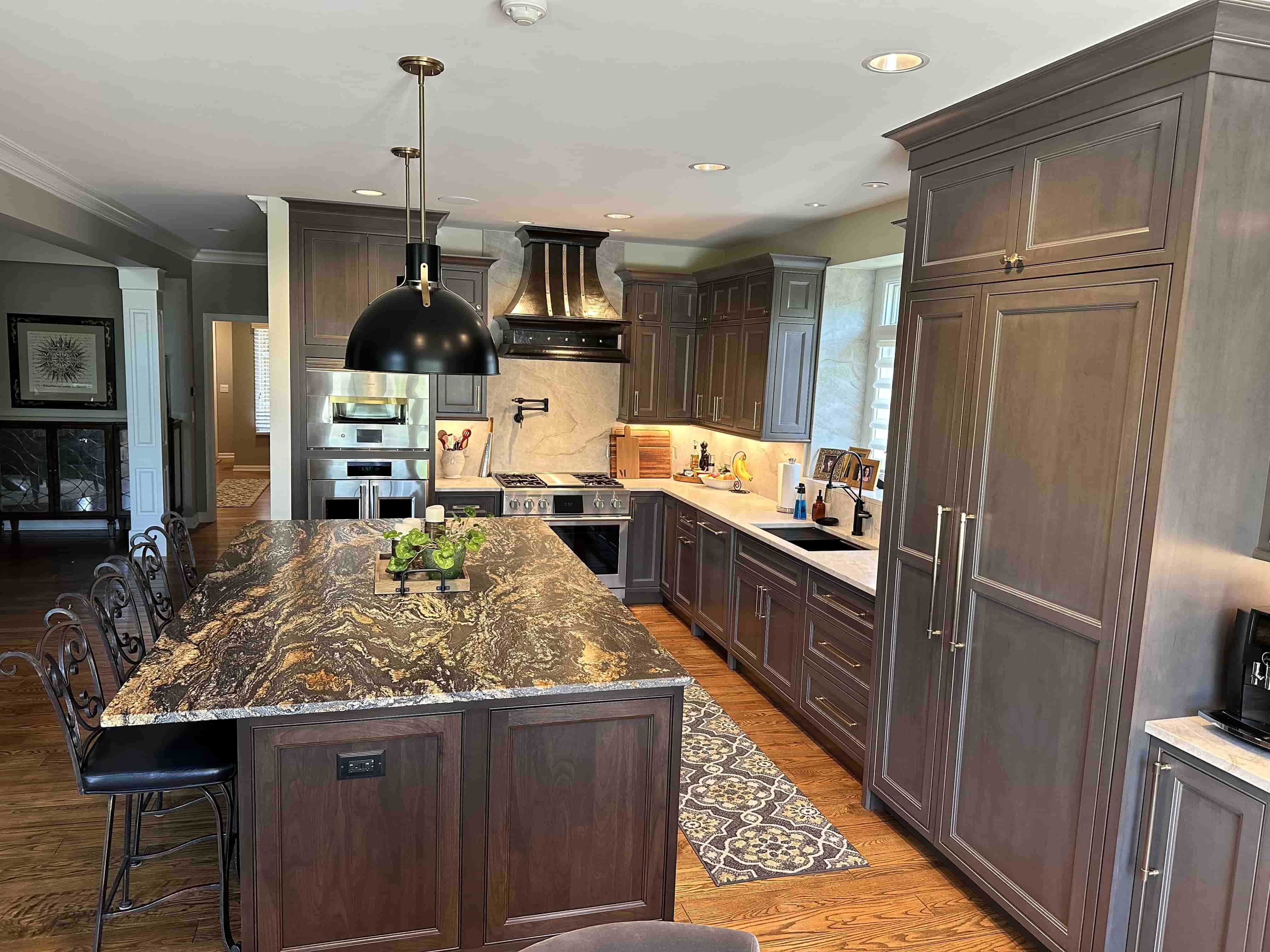 CopperSmith Signature SX11 Wall Mount Blackened Brass Range Hood with Brushed Stainless Steel Straps and Polished Brass Rivets in a cottage style kitchen with marble countertops wood cabinets and white marble backsplash