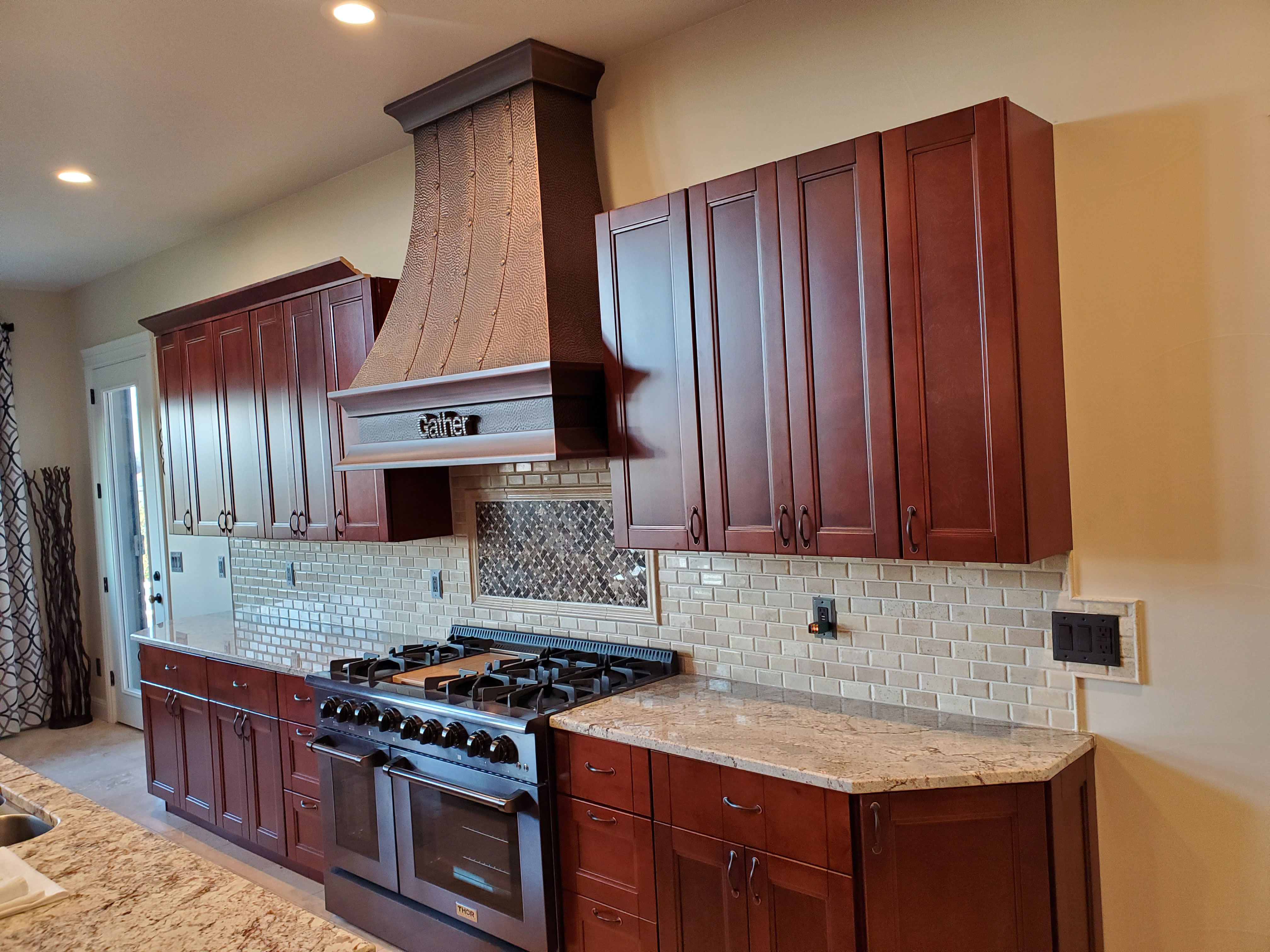 An industrial kitchen renovation featuring stylish range hood design, brown kitchen cabinets, marble countertops, brick backsplash