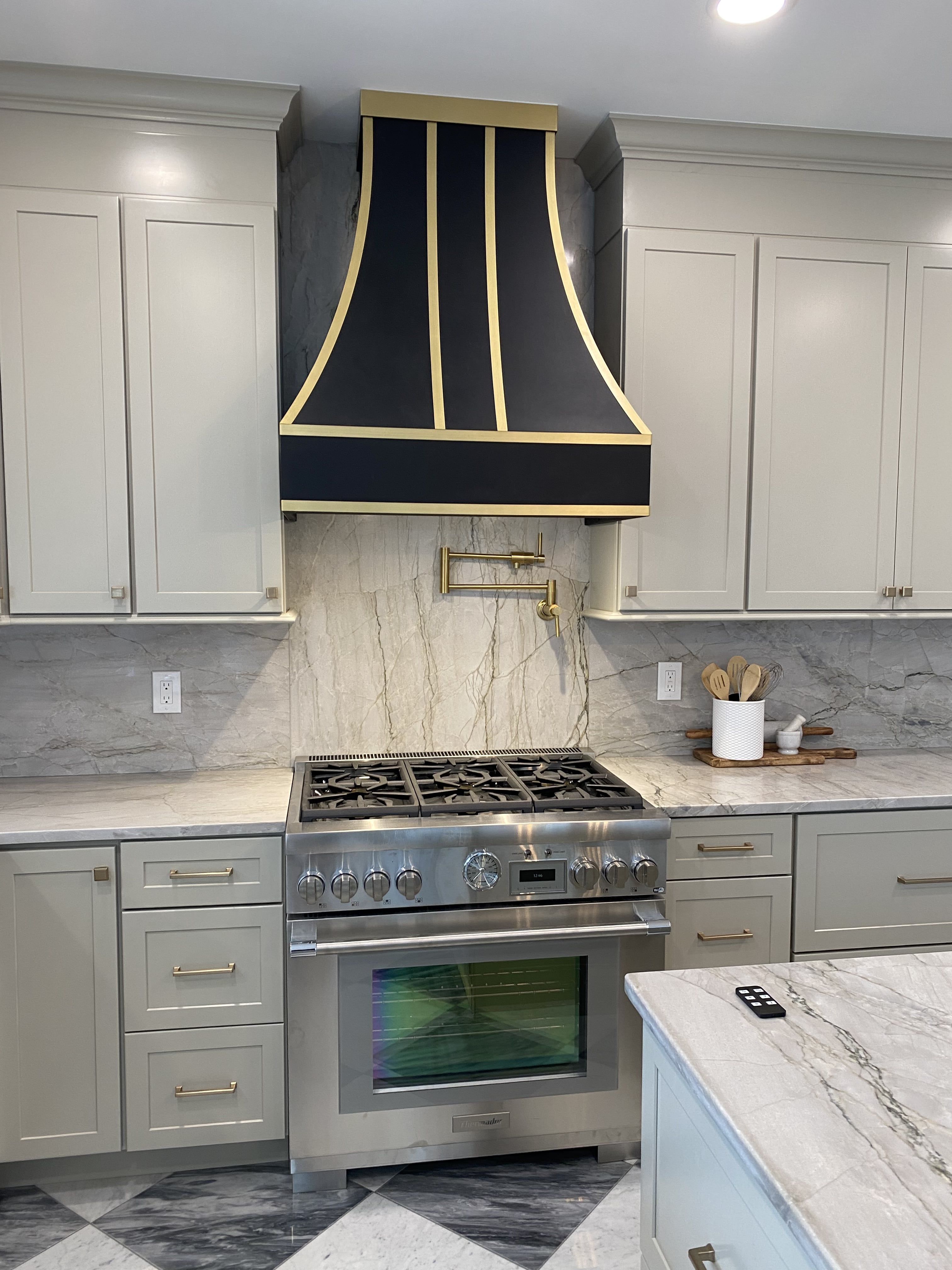 CopperSmith Signature SX4 Wall Mount Black Enamel Smooth Range Hood with Matte Brass Straps in a traditional style kitchen with Marble countertops marble tile backsplash and white cabinetry