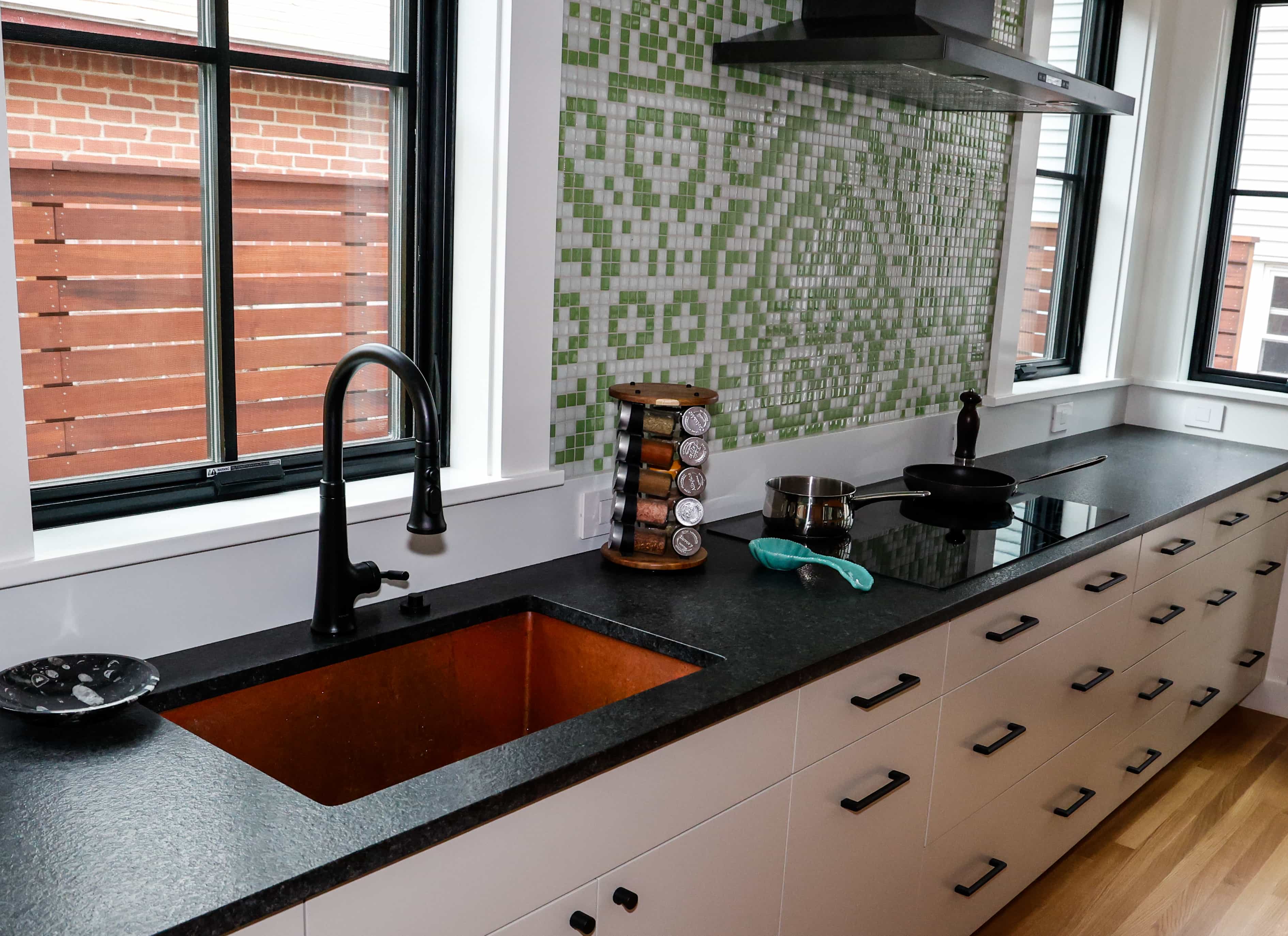 CopperSmith Undermount SB Weathered Copper Sink  in a contemporary kitchen with black countertops white cabinets and green and white tile backsplash